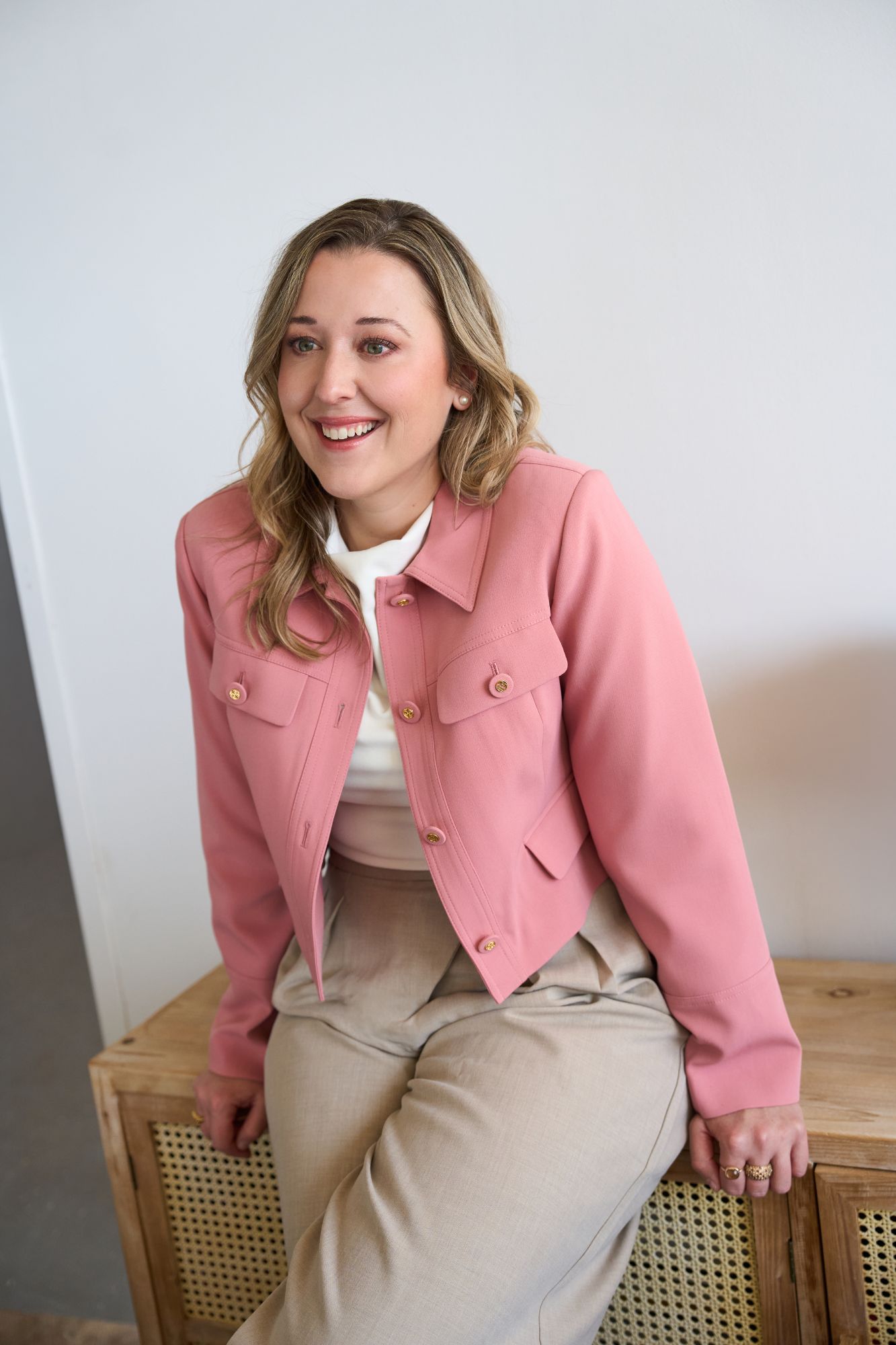 Founder of ELLA HR smiling on a wooden bench, dressed in a pink blazer over beige trousers.