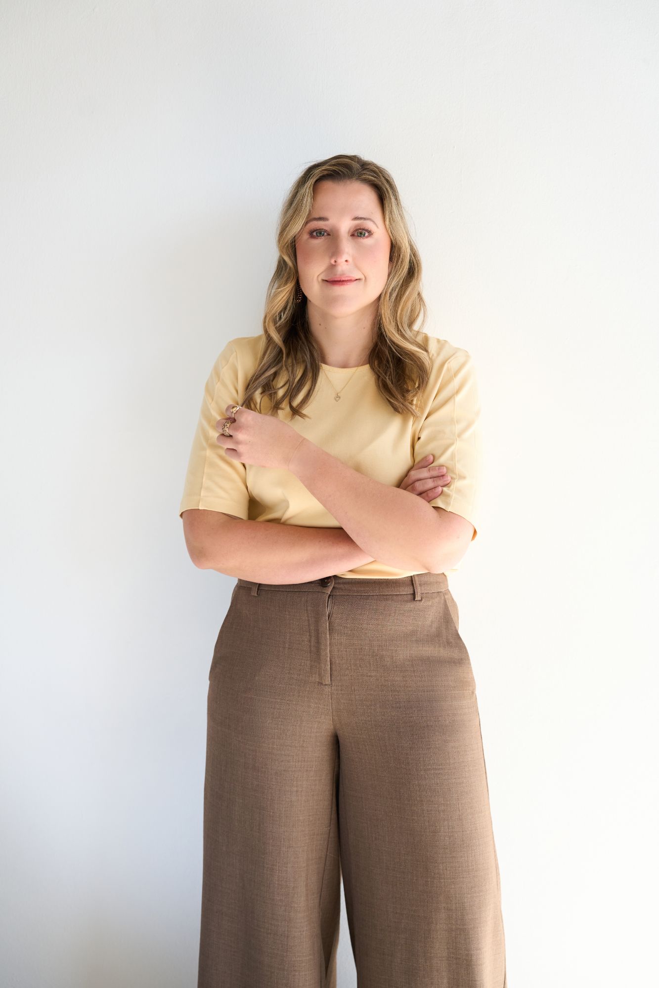 Founder of ELLA HR with arms crossed, dressed in a yellow shirt and brown trousers, against a white background.